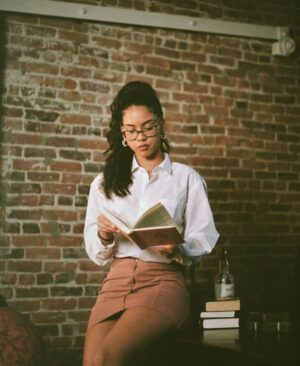 A young woman in a casual outfit reads a book indoors against a brick wall.